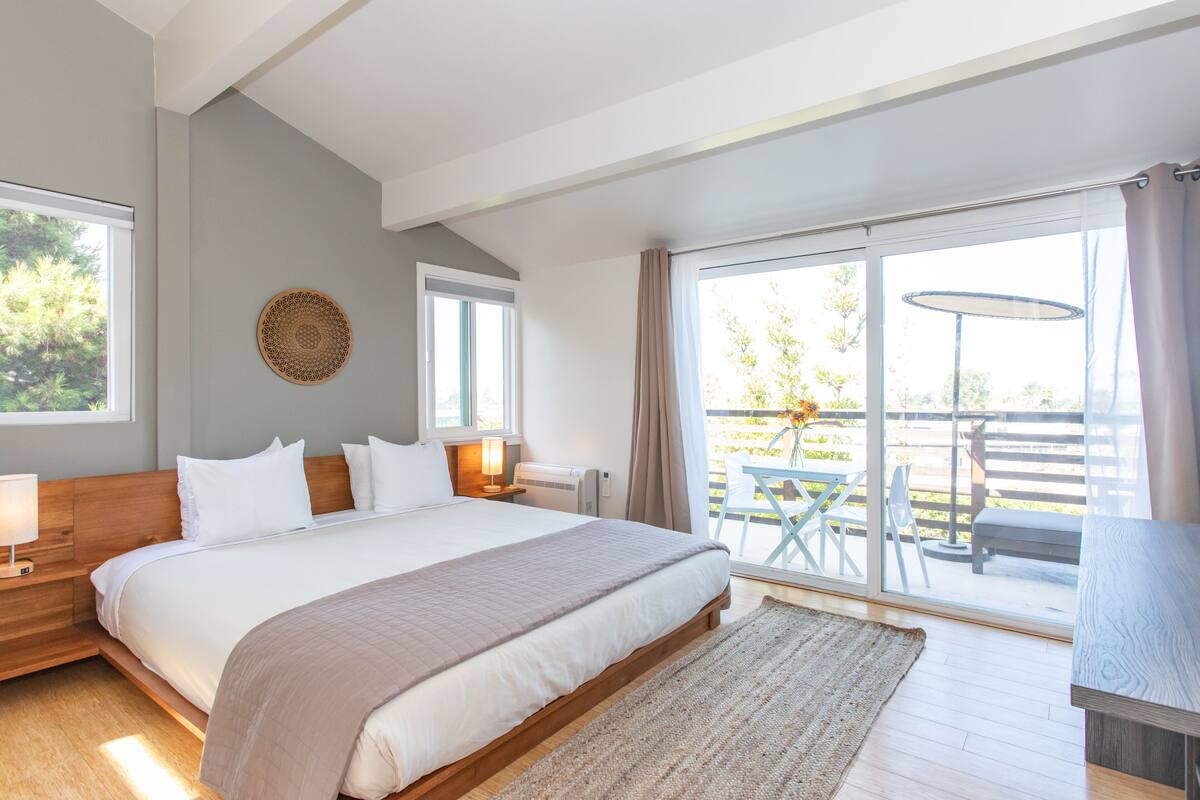 A bedroom with a large bed, a bedside table with a lamp, a wall hanging, and a view of an outdoor patio with a table and chairs.