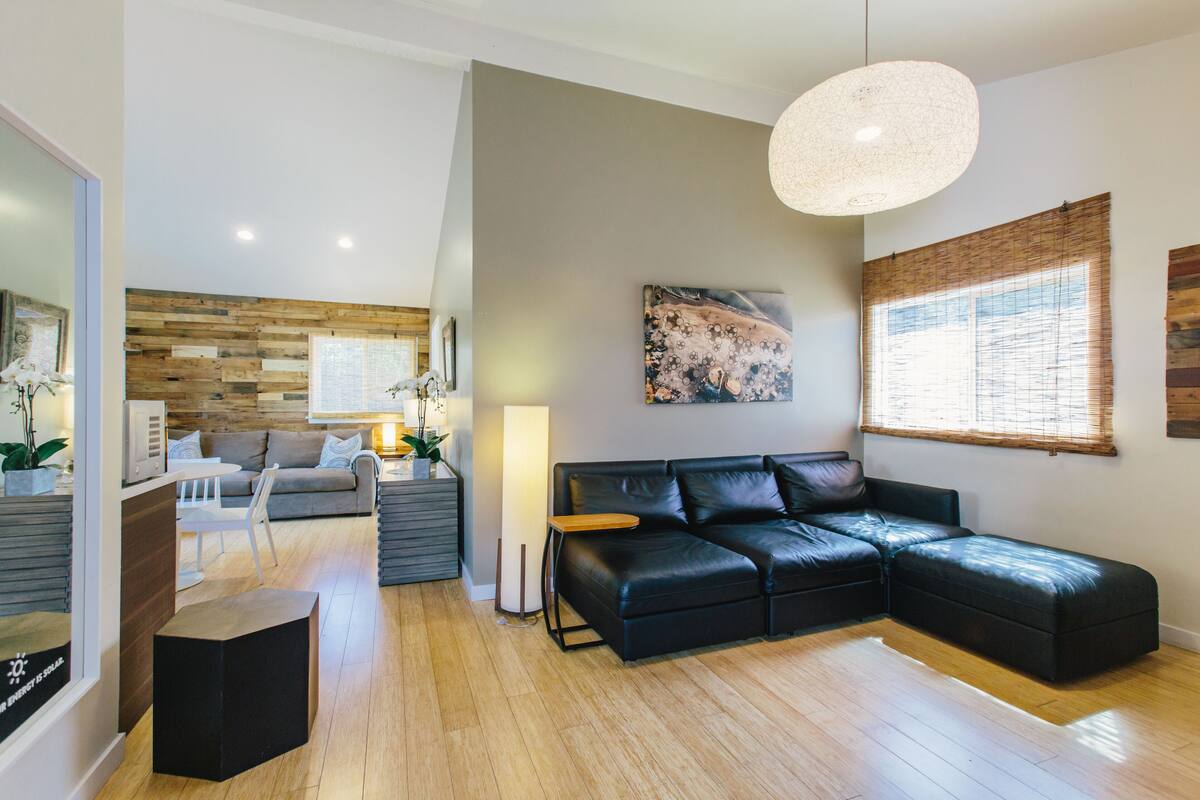A modern living room with a sectional sofa, a dining area with a white table and chairs, and a large window with bamboo blinds.
