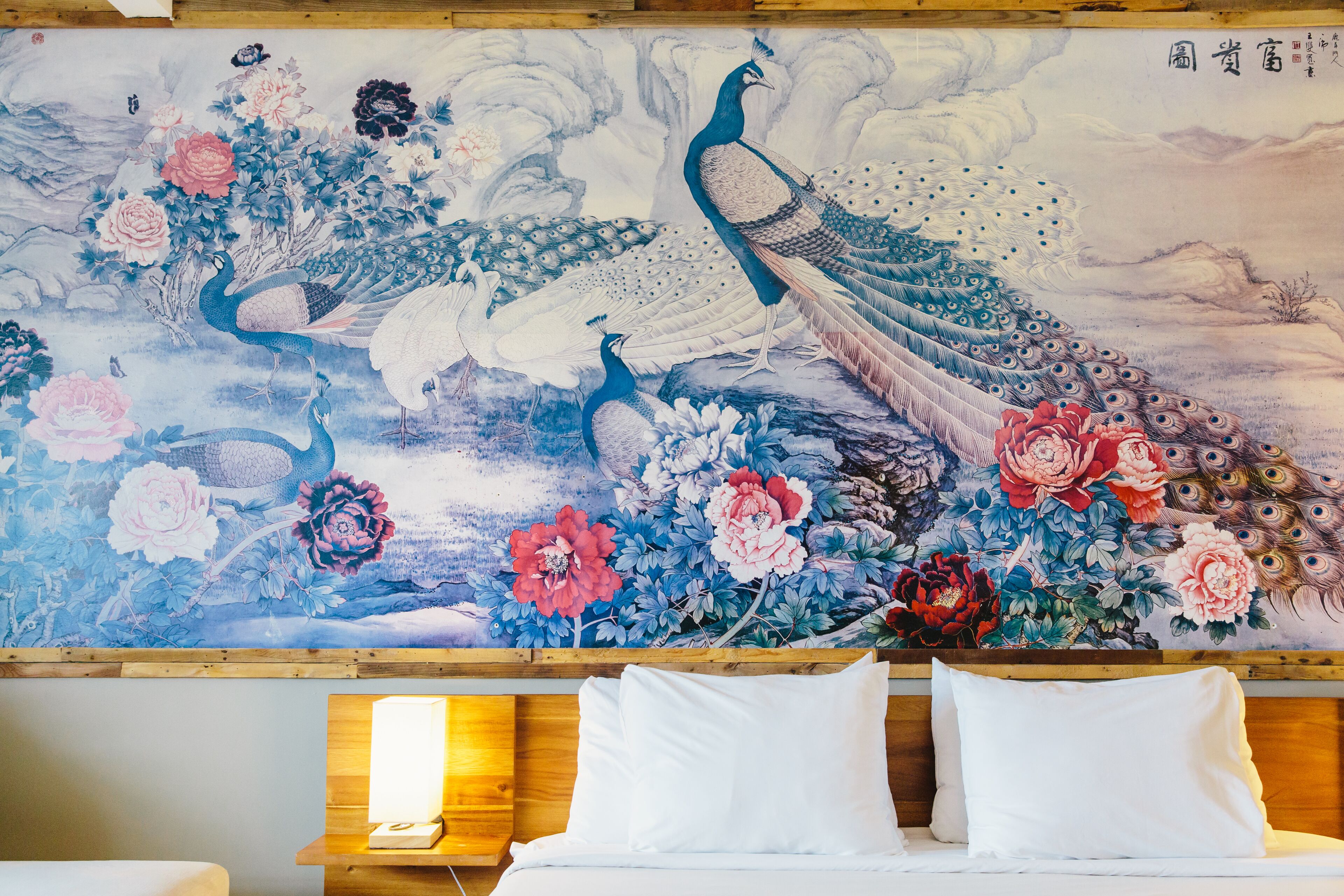 Un mural tradicional con pavos reales y flores en una pared, sobre una cama con almohadas blancas.