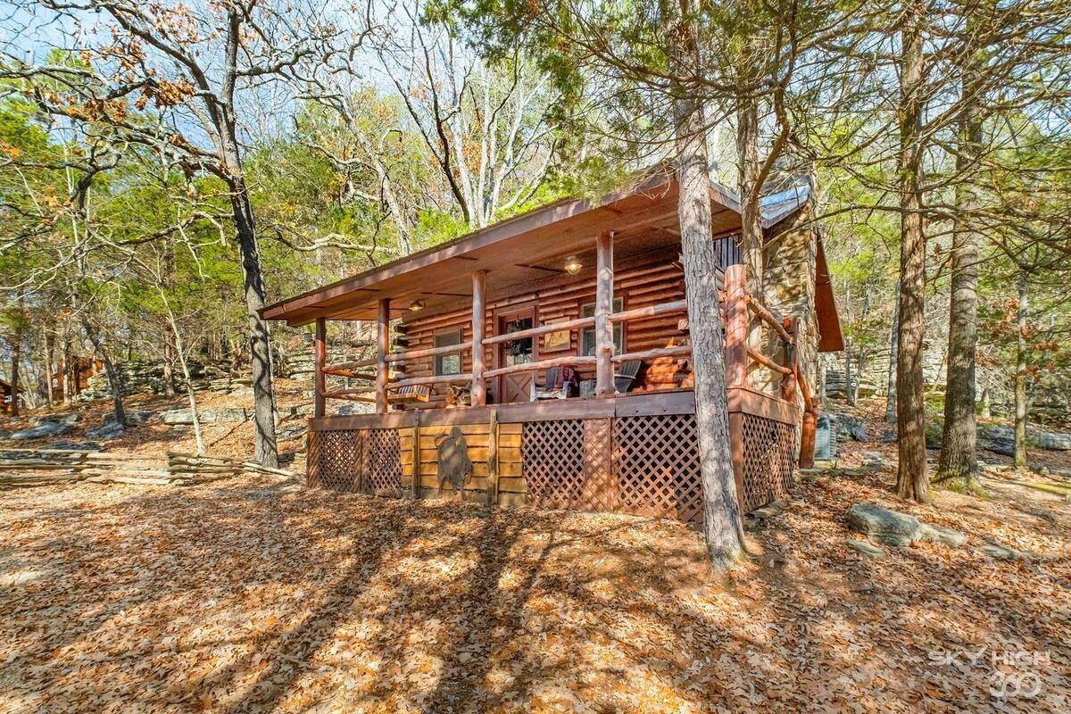 A rustic cabin with a wooden deck, surrounded by trees and a forest floor covered with leaves.