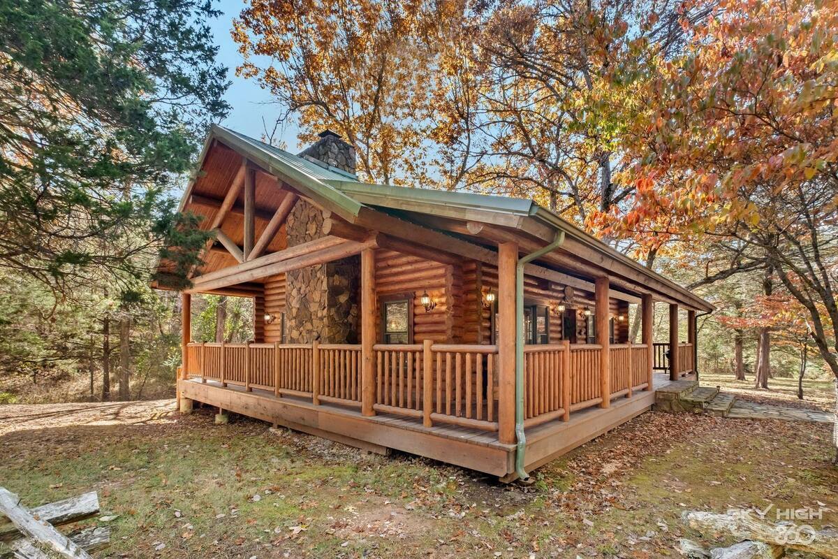 A rustic cabin with a covered porch and stone foundation, surrounded by autumn foliage.