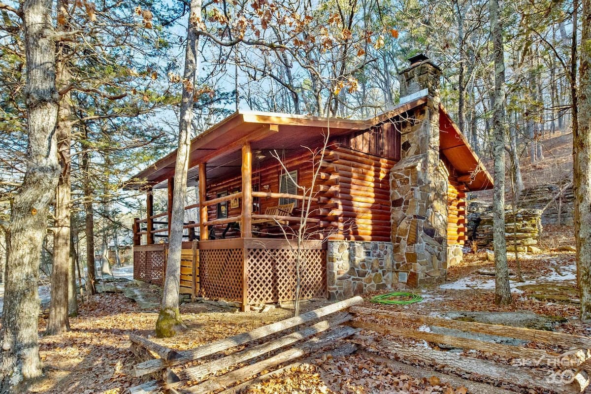 A log cabin with a stone foundation, surrounded by trees and a wooden deck.
