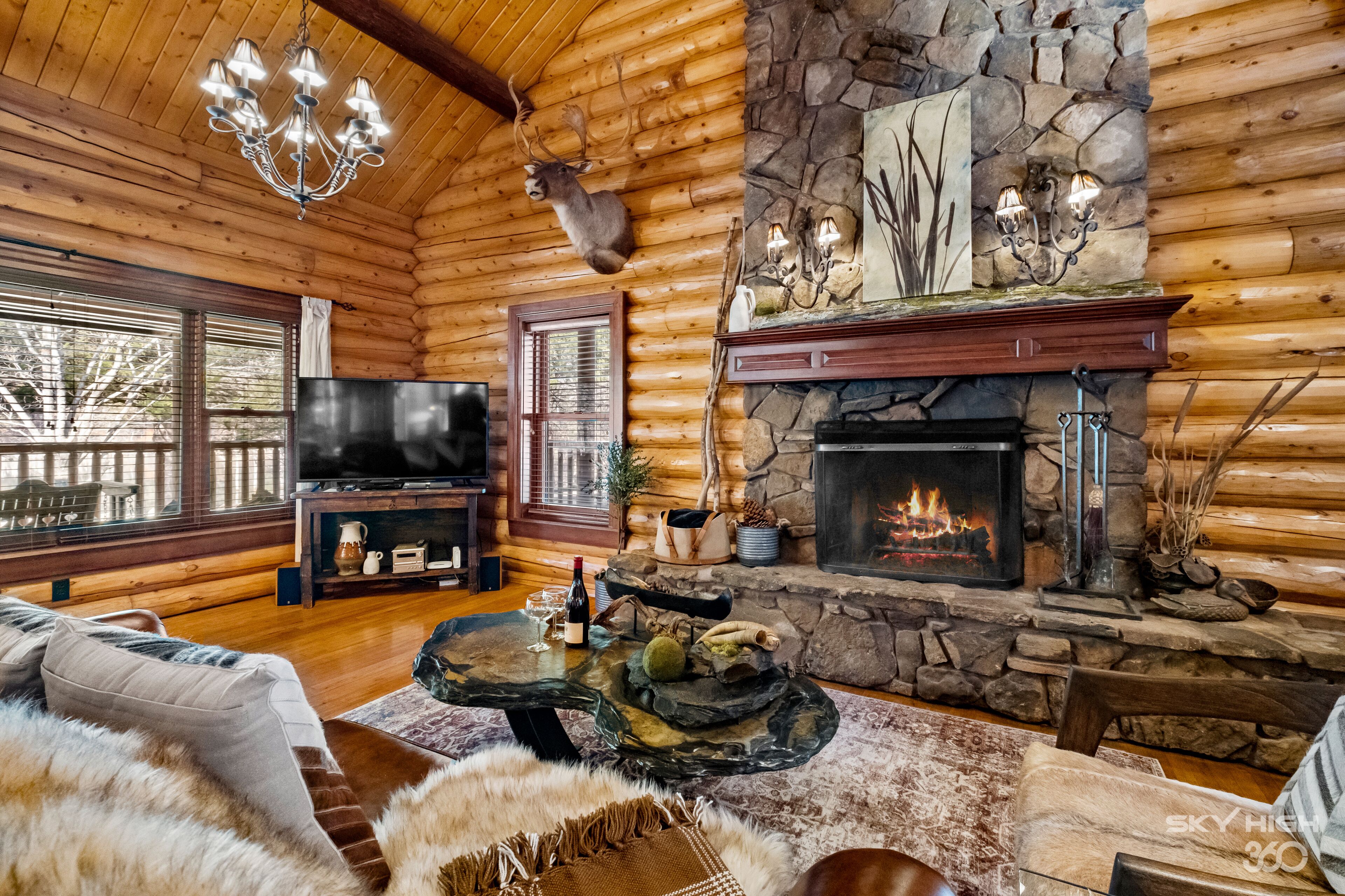 A log cabin interior with a stone fireplace, wooden walls, a chandelier, a television, and a cozy seating area.