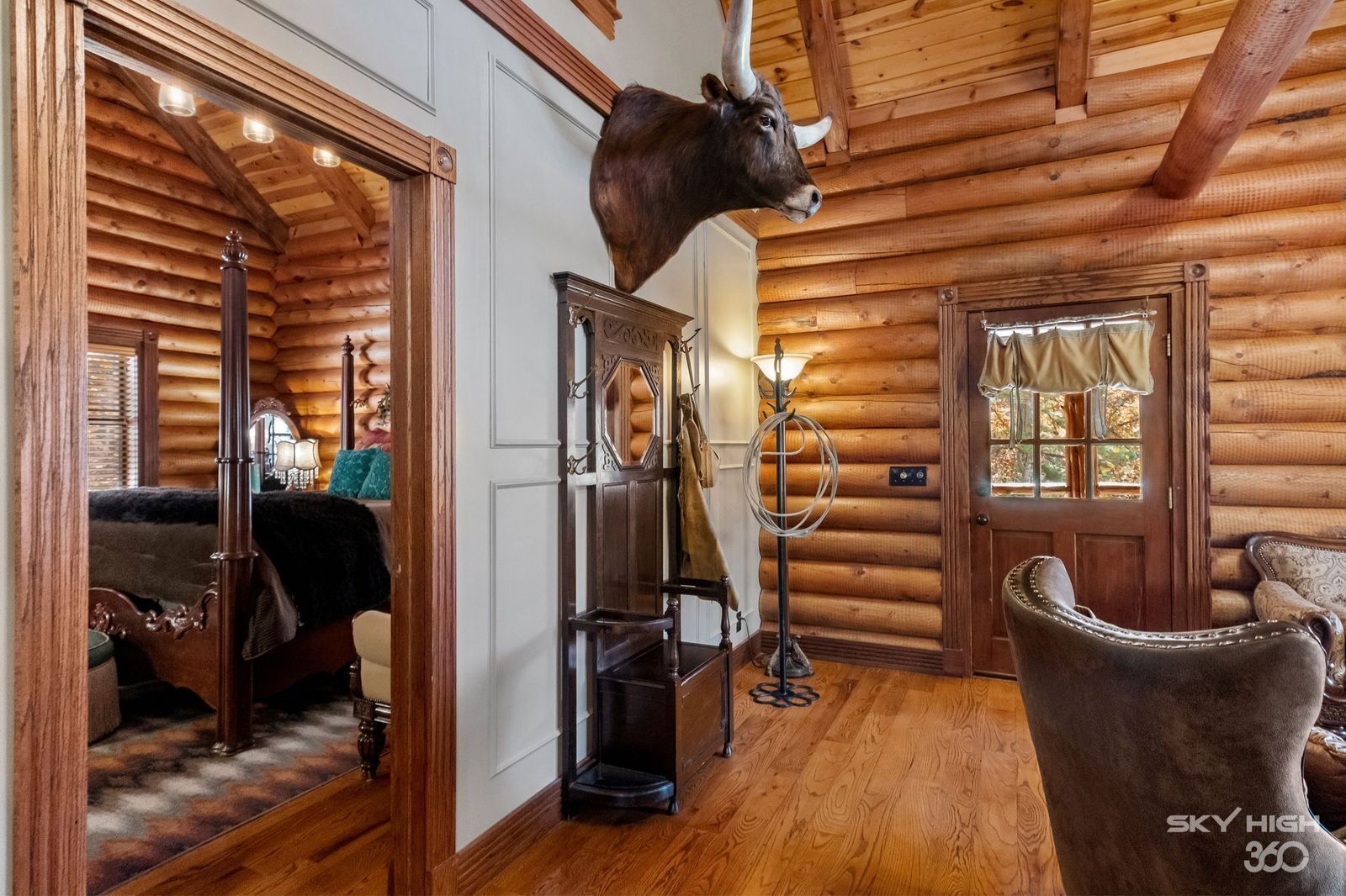 A log cabin interior with a bed, a chair, a lamp, and a mounted bull head.