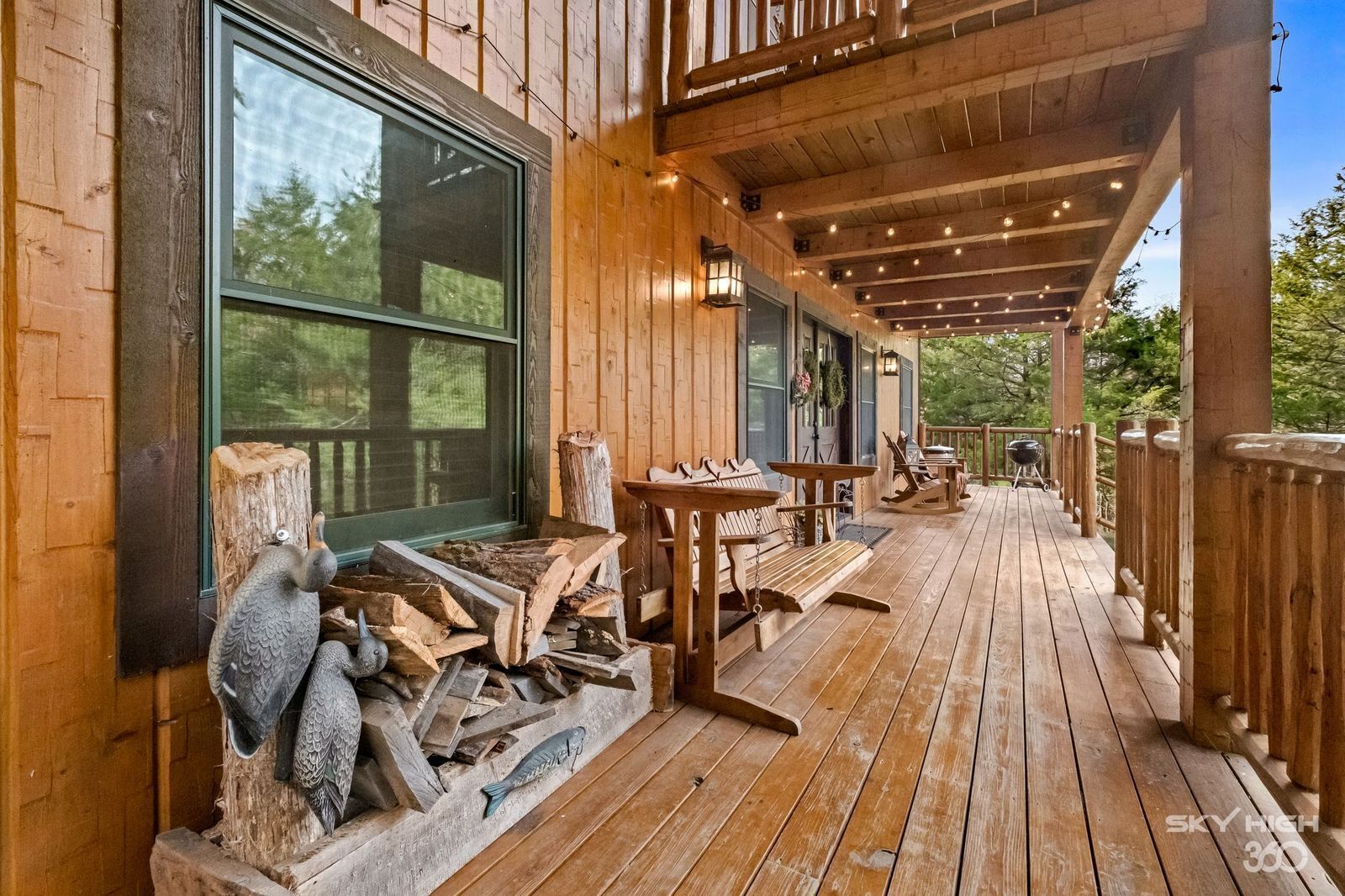 A wooden deck with outdoor seating, a large window, and a pile of firewood.