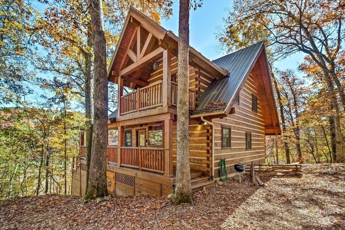 A log cabin with a balcony, a chimney, and a metal roof, surrounded by trees.
