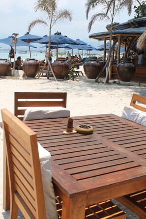 Ein Holztisch mit Stühlen auf einem Sandstrand, im Hintergrund Sonnenschirme und große Töpfe.