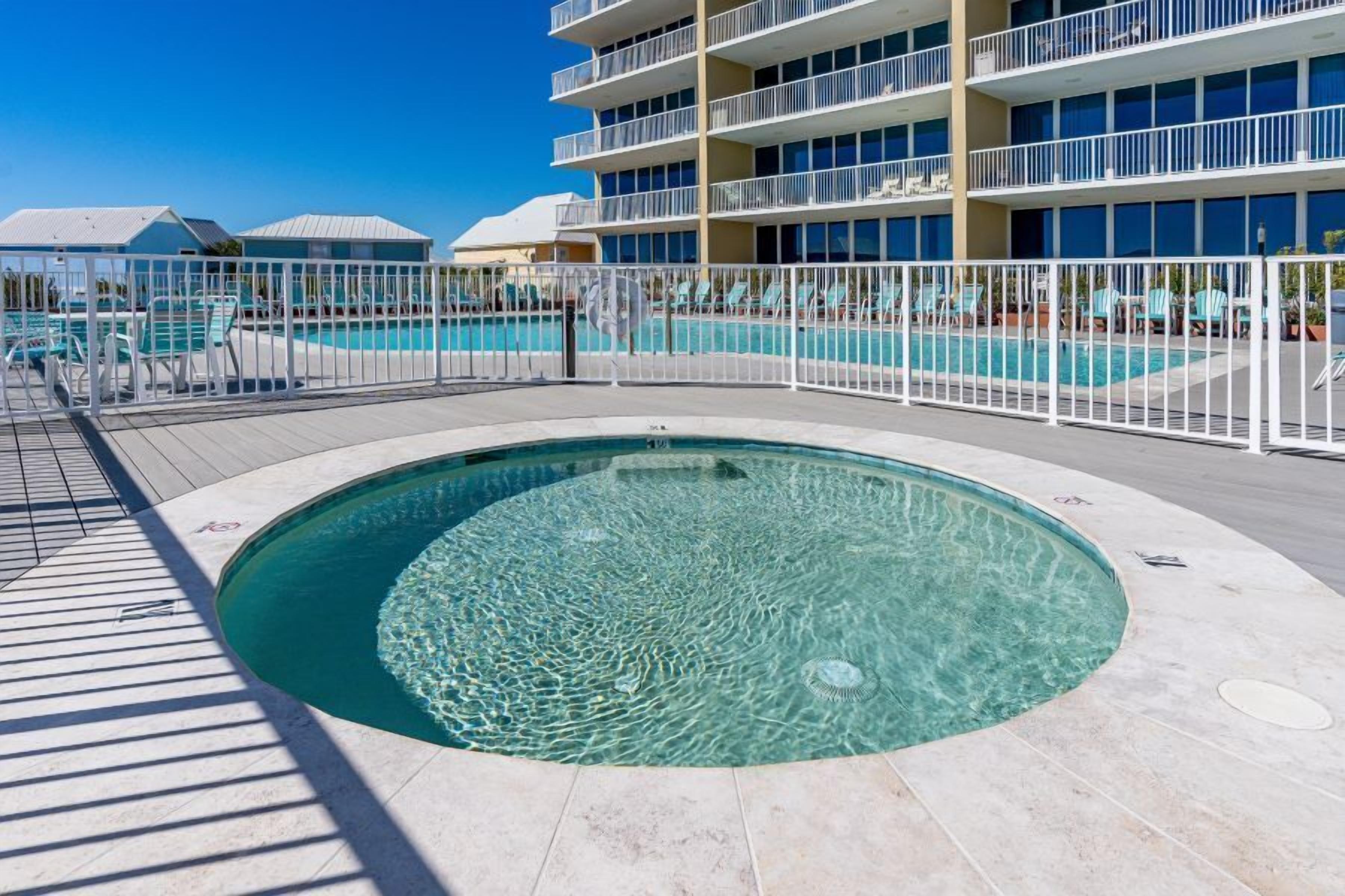 Uma área de piscina com piscina circular, cercada por uma grade branca, e um edifício com várias varandas ao fundo.