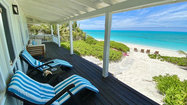 Une terrasse couverte avec deux chaises longues et une petite table, offrant une vue sur une plage de sable et un océan turquoise.