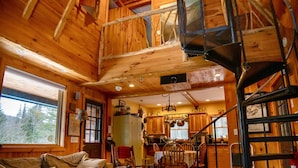 Een ruime houten chalet met hoge plafonds, een eethoek, een woonkamer met een bank en salontafel, en een spiltrap naar een verdieping erboven.