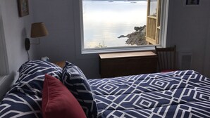 A bedroom with a large window overlooking a body of water, a bed with patterned bedding, a bedside lamp, and a small wooden table. - Secluded Oceanfrot cottage:Unbelievable oceanfront views-Screened in Gazebo (Wilsons Beach, Campobello Island)