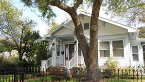 A two-story house with a front porch, a large tree in front, and a black metal fence. - Pet friendly cottage 1 block to beach fenced in yard with pergola & outdoor tub (Gulfport)