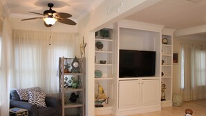 A living room with a ceiling fan, a TV on a white cabinet, a sofa with patterned cushions, and a bookshelf with decorative items. - Pet friendly cottage 1 block to beach fenced in yard with pergola & outdoor tub (Gulfport)