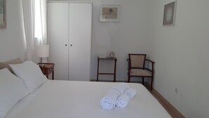 A neatly made bed with white linens and rolled towels, a bedside table with a lamp, a chair, and a wardrobe in a simple, clean room.