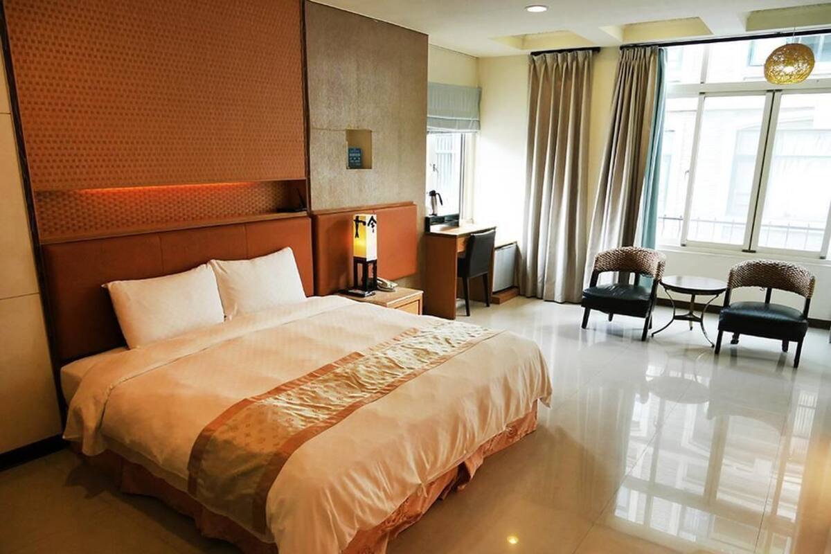 Habitación de hotel con una cama grande, un escritorio, dos sillas y vistas de la ciudad a través de amplias ventanas.