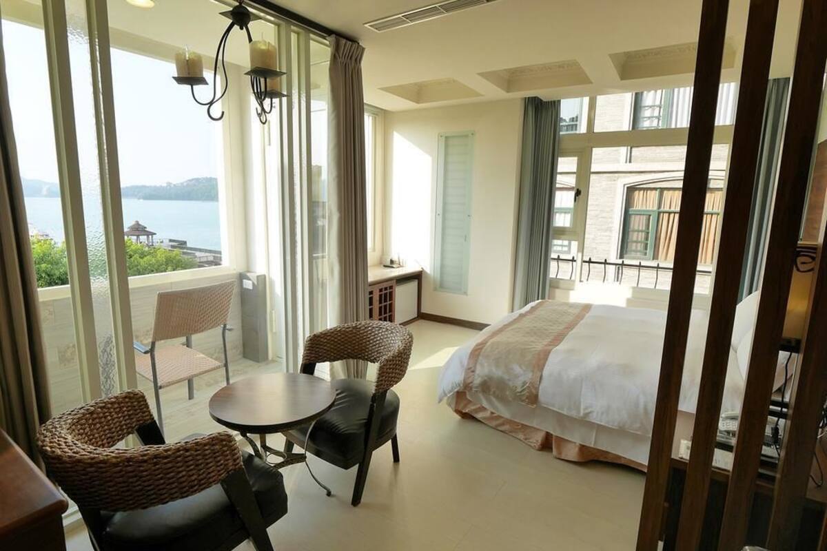 Habitación de hotel con cama, una mesa redonda, dos sillas de mimbre, vista al mar y un balcón con barandilla.
