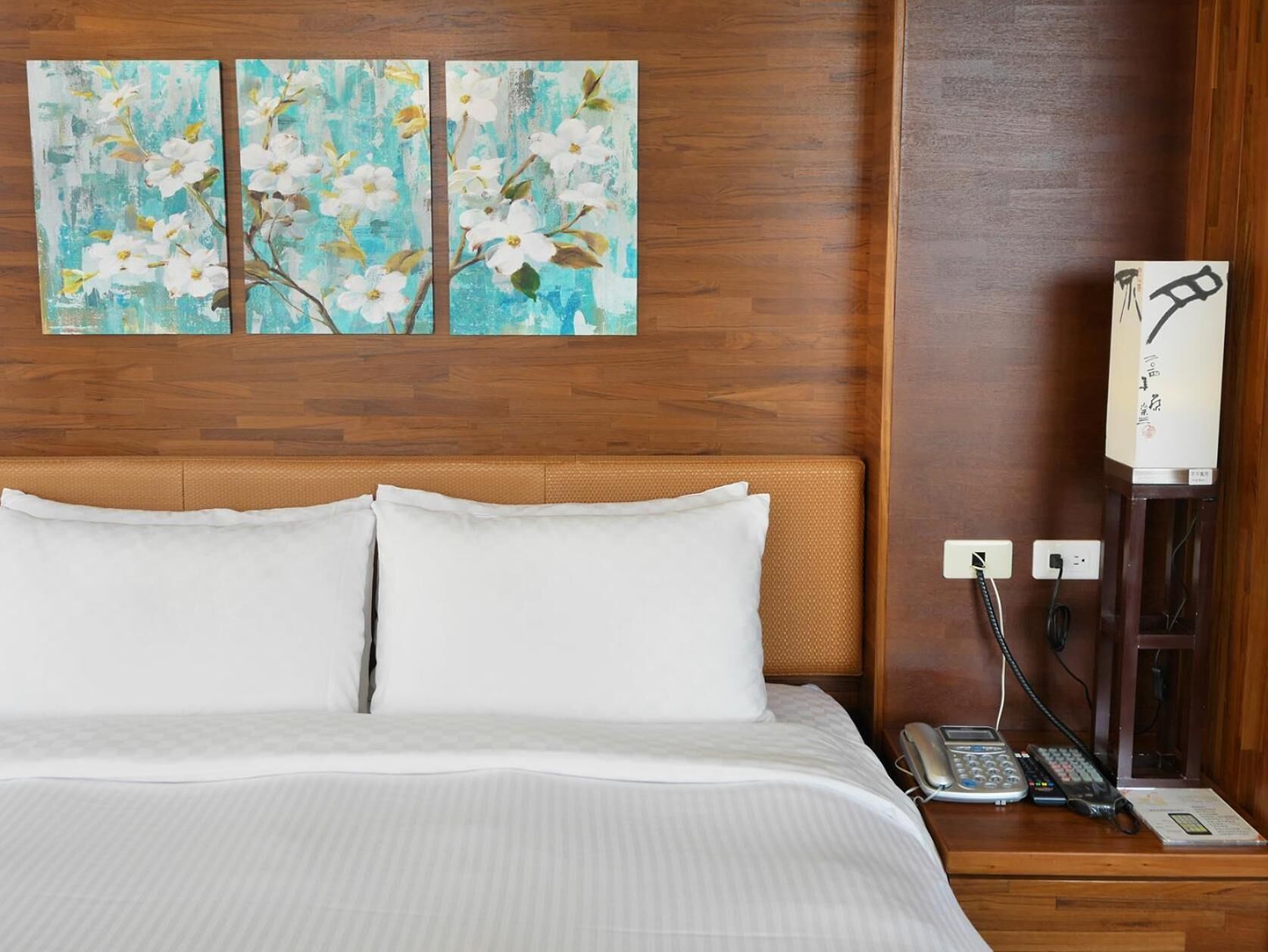 A hotel room with a bed, white pillows, a nightstand with a phone and a lamp, and two paintings of flowers on the wall.