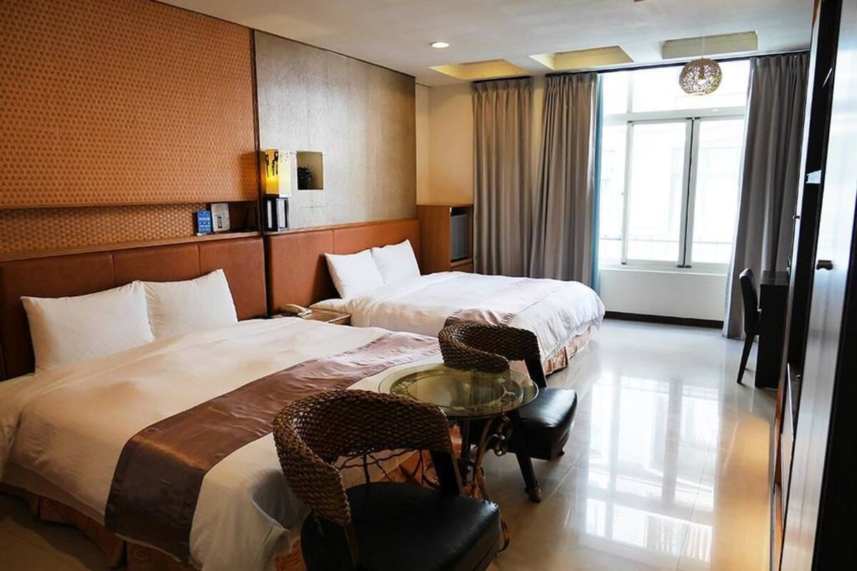 Habitación de hotel con dos camas, una mesa de vidrio, sillas de mimbre, televisor y un ventanal con cortinas.