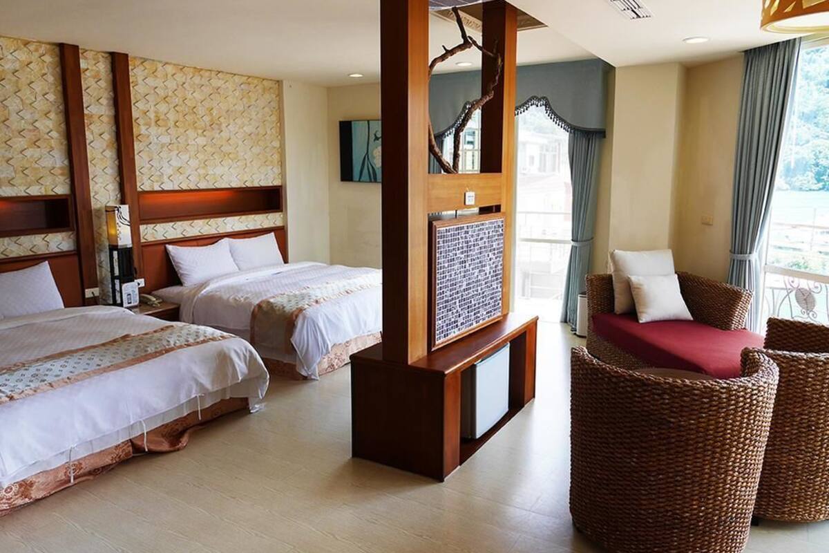 Habitación de hotel con dos camas, cabecera de madera, una silla de mimbre y vistas al exterior.