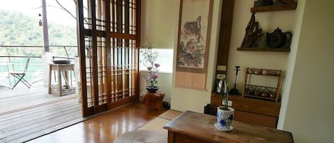 A traditional Japanese room with tatami flooring, a wooden table, a tatami mat, a sliding door, a view of a balcony, and a wall with a painting and shelves with pottery.