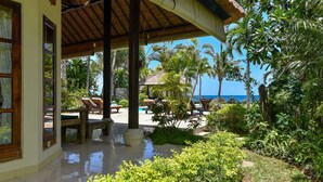 A covered outdoor area with lounge chairs and a pool, surrounded by lush greenery and a view of the ocean. - Modern, Luxury and Spacious Beach front Villa with Private Pool & Staff (Dencarik / Lovina)