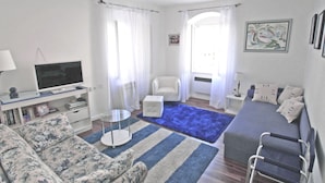 Ein Wohnzimmer mit einer Couch, einem Sessel und einem kleinen Tisch. Ein Fernseher steht auf einem Schrank und eine Bücherwand ist mit Büchern gefüllt.