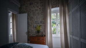 Un dormitorio con pared de piedra, un aparador de madera, una cama con almohadas estampadas y una ventana con cortinas.