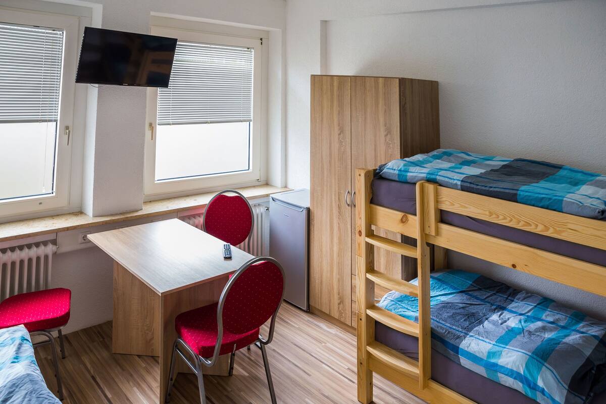 A dormitory room with bunk beds, a desk, a chair, a television, and a window with blinds.