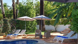 Une piscine entourée de chaises longues et de parasols, elle-même bordée d’une végétation luxuriante et d’une terrasse carrelée.