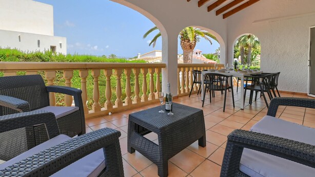 Un patio couvert aménagé avec des meubles en osier, une table avec deux verres, et une vue sur un bâtiment blanc et des palmiers.