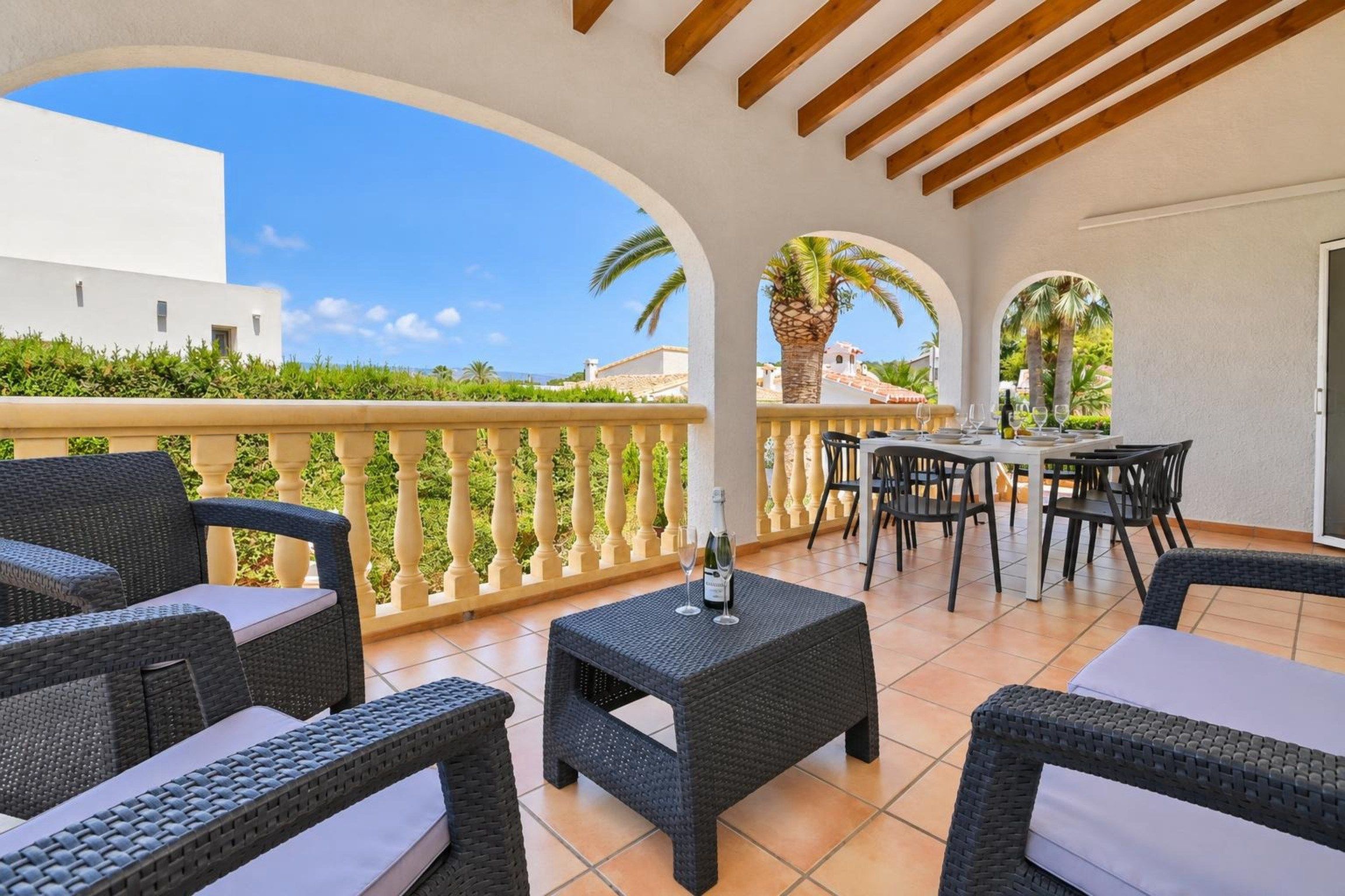 An outdoor patio with wicker furniture, a table set for dining, and a view of a white building and greenery.