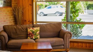 Een bruine bank met een bloemenkussen, een houten salontafel en een groot raam met uitzicht op een witte auto en begroeiing buiten.