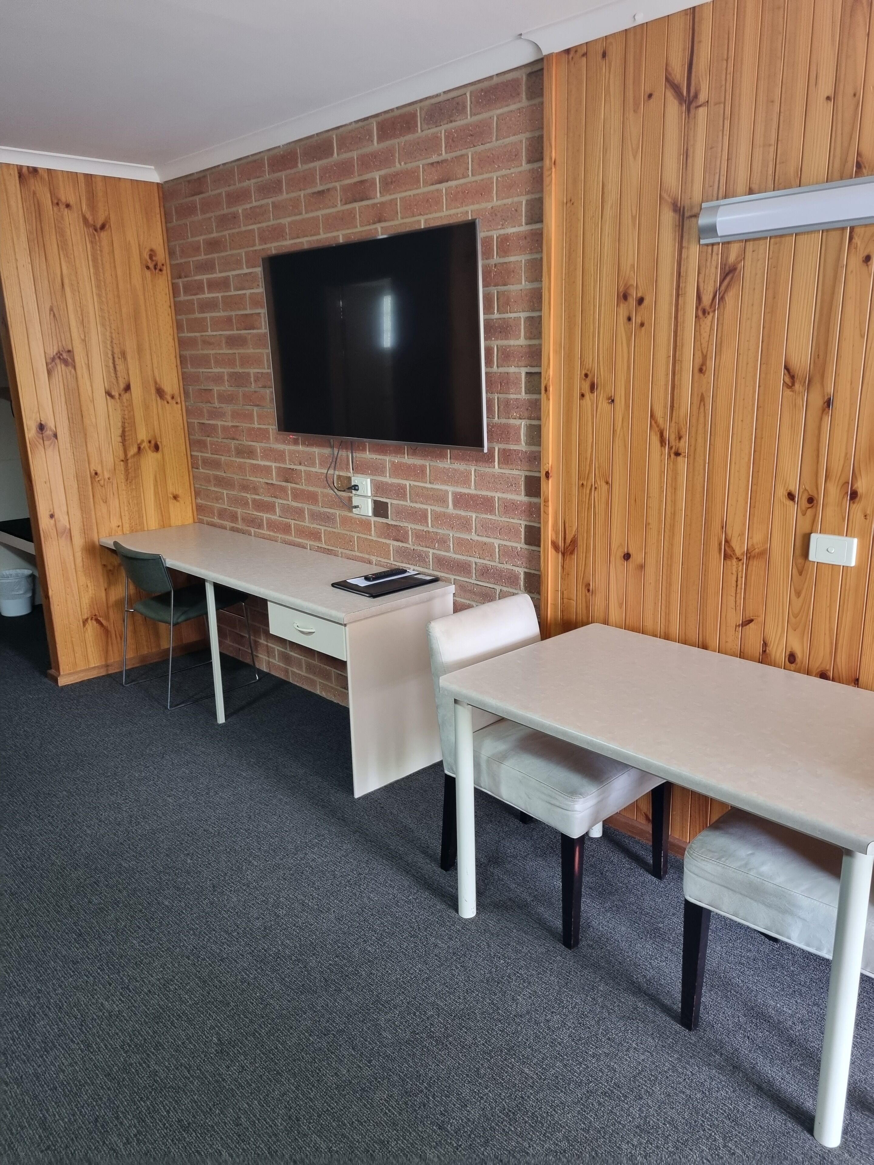 Een kamer met een bakstenen muur, een flatscreen televisie boven een bureau, een witte stoel en een houten wandpaneel.