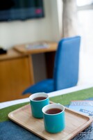 Two turquoise mugs on a wooden tray, a blue chair, and a wooden desk with a chair in a room with a window and a plant.