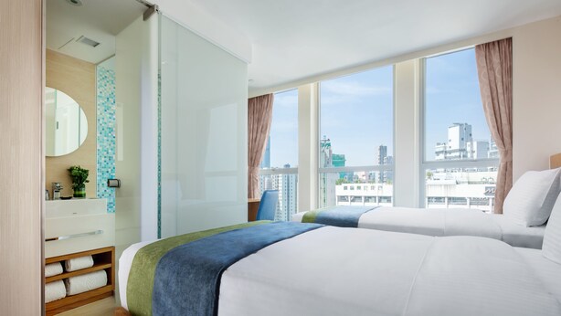 Habitación de hotel con dos camas, un ventanal grande, un baño con espejo y vistas a la ciudad.