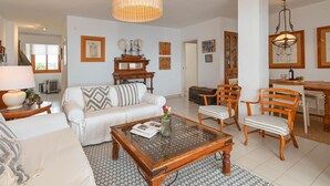 Ein geräumiges Wohnzimmer mit einem weißen Sofa, einem Glastisch und Holzstühlen. Es gibt eine Essecke mit Tisch und Stühlen, und ein großes Fenster lässt viel Tageslicht herein.