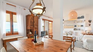 Ein Essbereich mit Holztisch, weißen Stühlen und einer Chandelierleuchte. Durch große Fenster ist der Meerblick sichtbar.