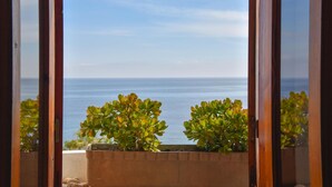 Blick durch ein Fenster mit Holzrahmen, das einen Balkon mit Topfpflanzen und einen klaren Meerblick zeigt.