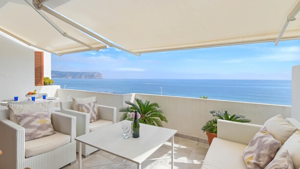 Ein Balkon mit weißem Tisch, einer weißen Couch und Korbstühlen mit Blick auf die Küste.