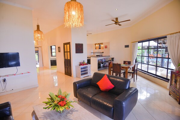 A spacious living room with a black leather sofa, a TV mounted on the wall, and a dining area with a table and chairs.