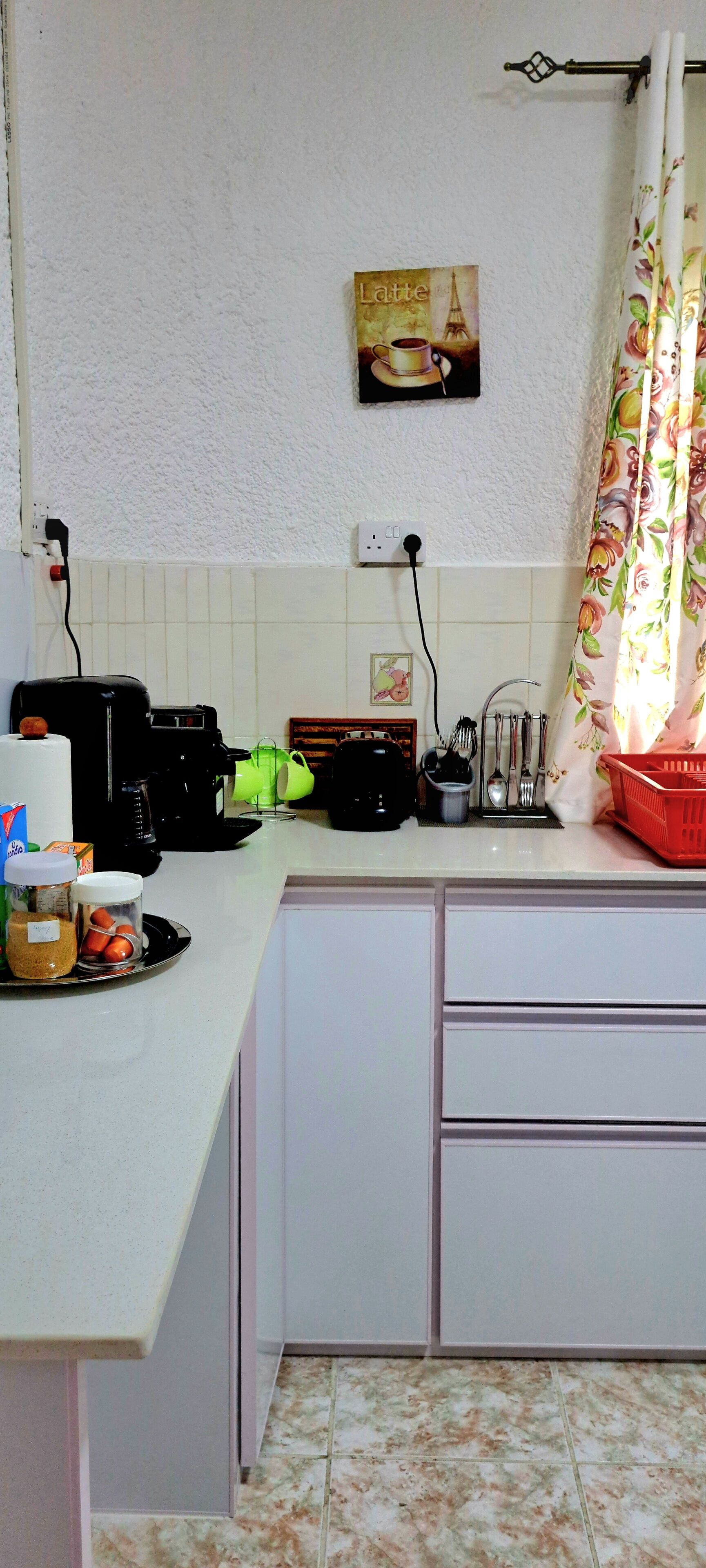 Una cocina con encimera blanca, cafetera, tostadora y hervidor. Hay un estante pequeño con frascos y una cesta.