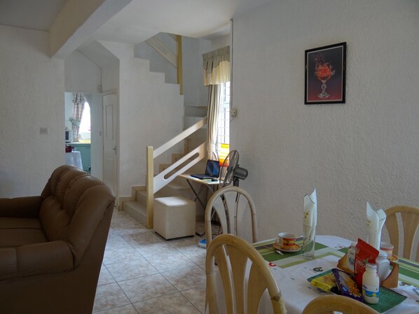 Ein Wohnzimmer mit braunem Sofa, einem Esstisch mit Stühlen, einer Treppe und einer Küche im Hintergrund.