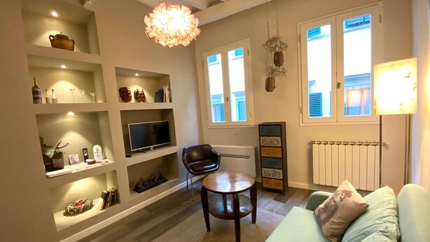 Una sala de estar con un sofá, televisión, estanterías con objetos decorativos, una silla y un radiador.