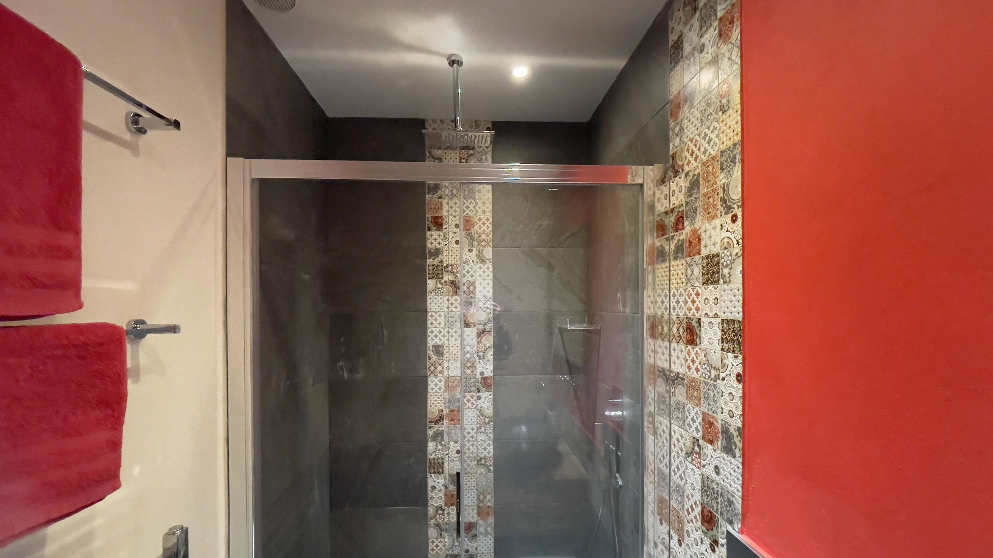A bathroom with a glass shower enclosure, a red towel rack, and patterned tiles on the walls.