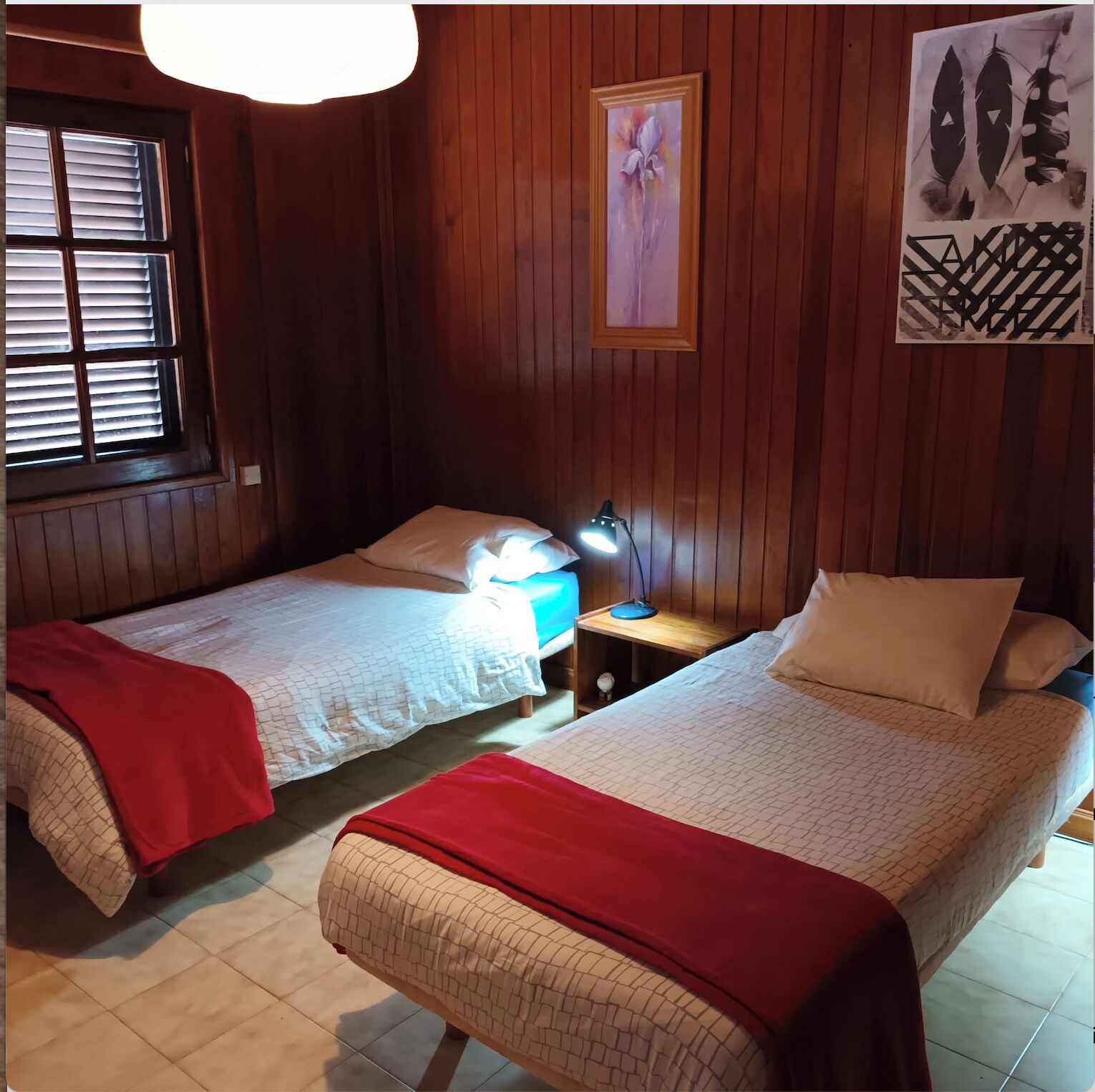 A room with two single beds, wooden walls, a window with blinds, a bedside table with a lamp, and artwork on the wall.