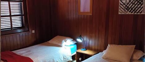 A room with two single beds, wooden walls, a window with blinds, a bedside table with a lamp, and artwork on the wall.