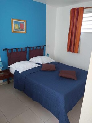 A bedroom with a blue wall, a bed with a blue bedspread and two pillows, a wooden nightstand with a lamp, and a painting on the wall. - Gîte Muskad, Creole charm and tropical calm at Cap Chevalier (Sainte Anne)
