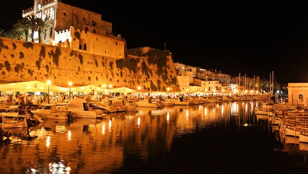 A historic waterfront area with a prominent castle-like structure, boats docked along the quay, and a bustling crowd of people.
