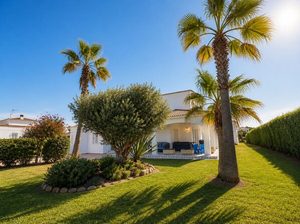 A white villa with a covered outdoor seating area, surrounded by well-maintained lawns and palm trees.