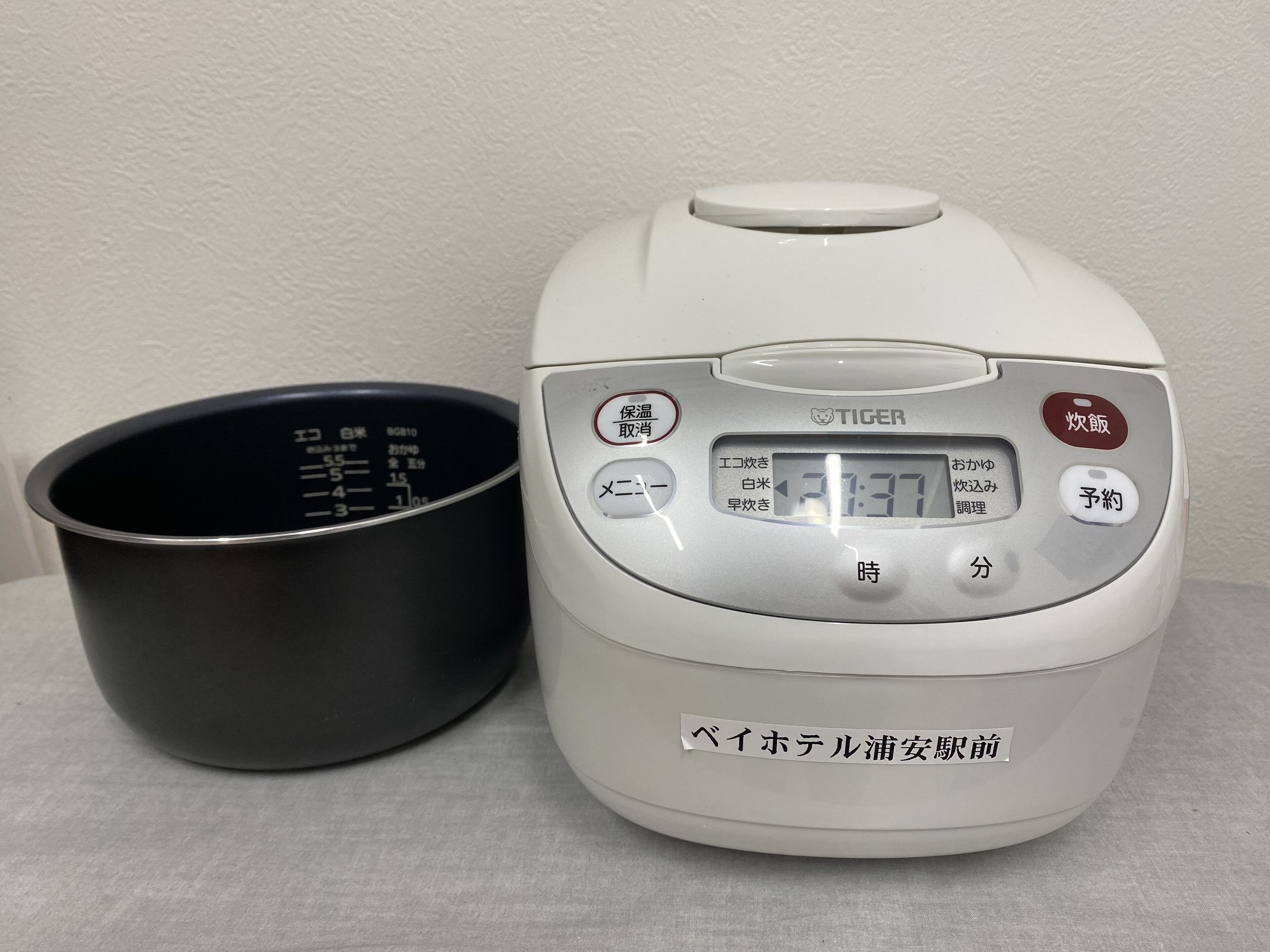 A white rice cooker with a digital display and buttons in Japanese, next to a black measuring cup.
